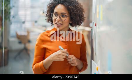Porträt einer schönen schwarzen Frau in eleganter Freizeitkleidung, die eine Präsentation in einem Besprechungsraum für ihre Kollegen macht. Weibliche Teamleiterin erklärt Daten und Statistiken mit einem Whiteboard und einem Marker Stockfoto