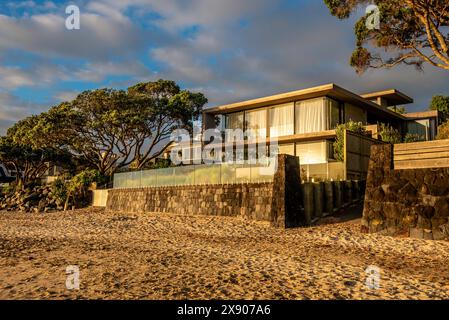 Die Sonne am frühen Morgen spiegelt sich von den Fenstern eines modernen Hauses am Strand in Campbells Bay, Neuseeland Stockfoto