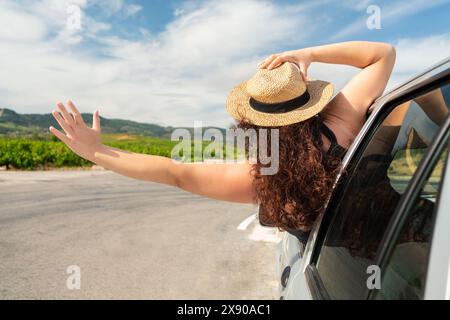 Rückfahraufnahme einer jungen Frau, die sich aus ihrem Autofenster beugt, mit ausgestreckten Armen auf der Straße Stockfoto