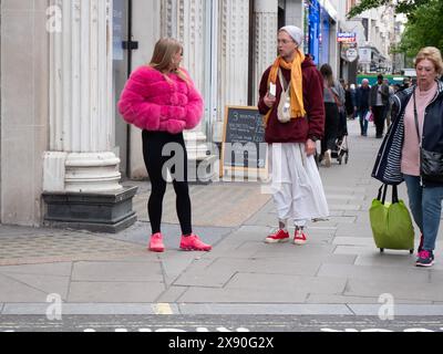 Hare Krishna, Follower, der Bücher an Käufer verteilt, spricht mit einer jungen modischen Frau in rosa Jacke und Schuhen in der Oxford Street, London, Großbritannien Stockfoto