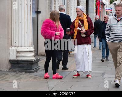 Hare Krishna, Follower, der Bücher an Käufer verteilt, spricht mit einer jungen modischen Frau in rosa Jacke und Schuhen in der Oxford Street, London, Großbritannien Stockfoto