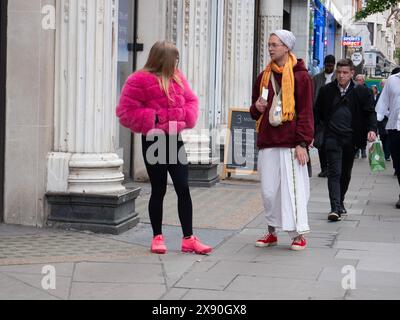 Hare Krishna, Follower, der Bücher an Käufer verteilt, spricht mit einer jungen modischen Frau in rosa Jacke und Schuhen in der Oxford Street, London, Großbritannien Stockfoto