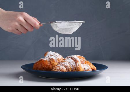 Frau mit einem Sieb, das Puderzucker auf Croissants streut, am weißen Holztisch, Nahaufnahme Stockfoto