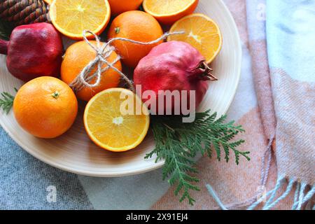 Ein Holzteller mit frischen Orangen und Granatäpfeln, einige in Scheiben geschnitten, garniert mit einem Tannenzweig und Garn auf einer pastellfarbenen Serviette Stockfoto