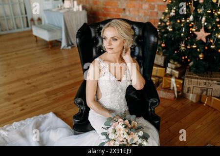 Braut in einem weißen Hochzeitskleid mit Blumenstrauß in einem Ledersessel in der Nähe eines Weihnachtsbaums und präsentiert sich in einer gemütlichen Inneneinrichtung Stockfoto