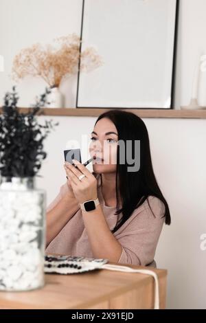 Eine Frau mit schwarzem Haar trägt Lippenstift vor einem kleinen Spiegel in einer legeren, gemütlichen Atmosphäre mit minimalistischem Dekor auf Stockfoto