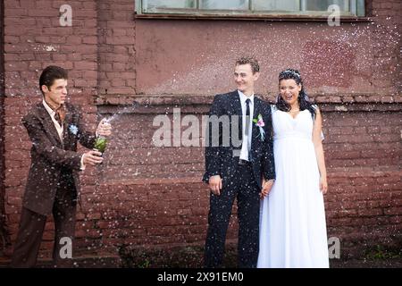 Ein Paar feiert ihre Hochzeit mit einem Freund, der eine Champagnerflasche platzt, alle stehen vor einer Ziegelmauer, Weißrussland, Minsk Stockfoto