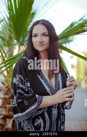 Eine Frau mit dunklen Haaren in einem schwarzen Kleid steht draußen neben einer Palme, sieht entspannt und lächelnd aus, Truthahn, Antalya Stockfoto