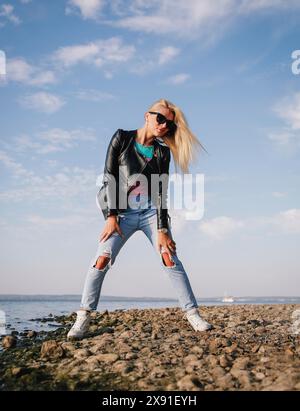 Eine blonde Frau in Lederjacke, zerrissener Jeans und Sonnenbrille steht an einem felsigen Ufer unter blauem Himmel mit verstreuten Wolken, Weißrussland, Minsk Stockfoto