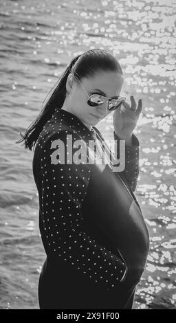 Schwangere Frau im Schwarzweißfoto, mit Sonnenbrille und Polka Dot Blazer, stehend am sonnendurchfluteten Meer Wasser. Weißrussland, Minsk Stockfoto