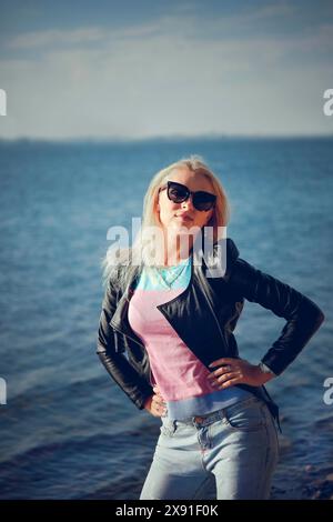 Blonde Frau in Sonnenbrille und Lederjacke steht selbstbewusst am Meer an einem sonnigen Tag, Weißrussland, Minsk Stockfoto