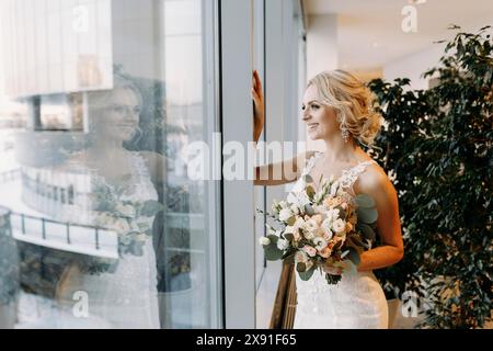 Die Braut in einem weißen Hochzeitskleid mit einem Blumenstrauß blickt aus einem großen Fenster, das ihr Bild reflektiert, in einem modernen Gebäude mit Grün im Inneren Stockfoto