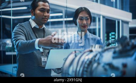 Ein junges Team multikultureller Ingenieure verwendet Blaupausen, Tablet- und Laptopcomputer, analysiert und diskutiert, wie ein moderner Elektroturbinenmotor funktioniert. High-Tech-Forschungslabor mit moderner Ausrüstung. Stockfoto