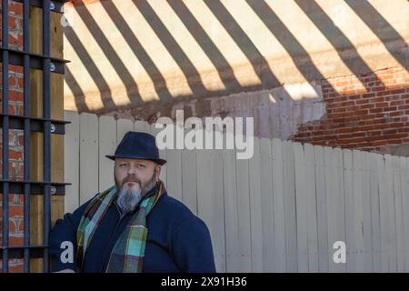 Mit Hut, kariertem Schal und Pfeife lehnt sich ein Mann an eine alte rustikale Gebäudewand an einem Fenster. Stockfoto