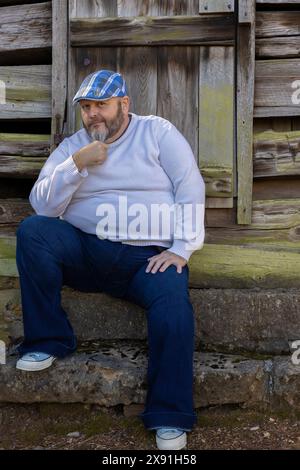 Ein Mann mit karierter blauer Mütze, weißem Pullover, Jeans und Tennisschuhen mit Bart posiert vor einem alten rustikalen Gebäude. Stockfoto