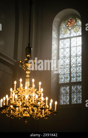 Große Aufnahme eines großen Fensters und eines antiken Kronleuchters in einer großen Kirche. Wunderschönes Buntglas, das die Sonne im Inneren des Gebäudes erleuchtet, und Kerzen, die ein warmes Leuchten ausstrahlen. Friedliche Atmosphäre Stockfoto