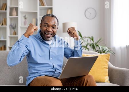 Aufgeregter Mann in blauem Hemd, der Erfolg mit erhobenen Fäusten feiert, zu Hause auf einer Couch mit einem Laptop sitzt. Sieg, Glück und Leistung in einem hellen, modernen Wohnzimmer. Stockfoto