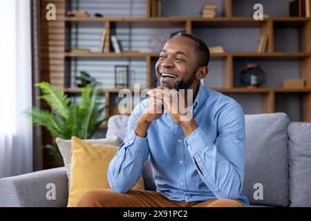 Eine glückliche Person in einem blauen Hemd lacht und entspannt auf einer gemütlichen Couch, umgeben von einem stilvollen, modernen Wohnzimmer mit einem Bücherregal im Hintergrund und einer grünen Pflanze in der Nähe. Stockfoto