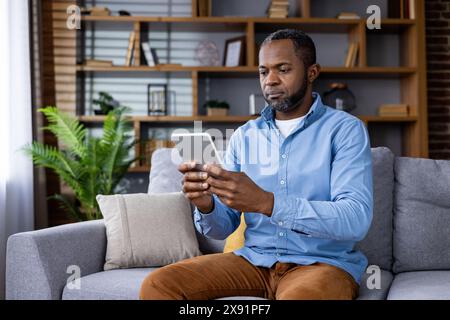 In einem modernen Wohnzimmer sitzt ein Mann auf einer Couch mit einem Tablet. Die Einrichtung und die Regale verstärken das moderne Ambiente und schaffen eine gemütliche Umgebung für Arbeit oder Entspannung Stockfoto
