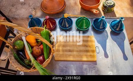 Ein lebendiger Korb mit frischem Gemüse neben farbenfrohen Tagine-Töpfen auf einem hölzernen Schneidebrett in einer rustikalen Außenküche. Stockfoto