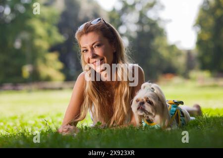 Eine fröhliche junge Frau liegt auf Gras in einem sonnendurchfluteten Park und genießt einen friedlichen Moment mit ihrem liebenswerten Shih Tzu Hund. Stockfoto