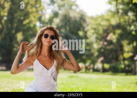 Eine junge Frau mit Sonnenbrille und weißem Kleid genießt einen unbeschwerten Moment und spielt mit ihren Haaren in einem sonnendurchfluteten Park, umgeben von Bäumen. Stockfoto