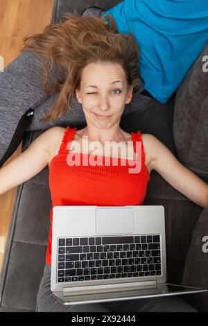 Eine fröhliche junge Frau in einem roten Oberteil legt sich auf eine graue Couch, benutzt einen Laptop und lächelt in die Kamera, zeigt Komfort und lässigen Lebensstil. Stockfoto