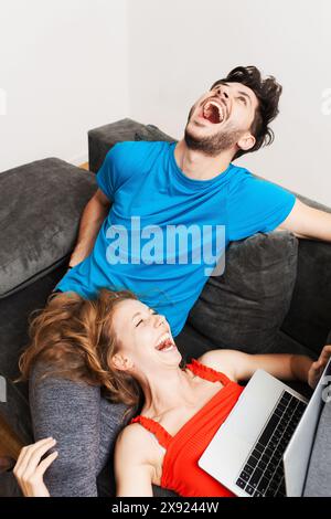 Ein glückliches junges Paar genießt einen urkomischen Moment, während es sich auf einem Sofa entspannt und über etwas amüsantes auf seinem Laptop lacht. Stockfoto