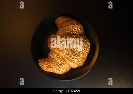 Ein frisch gebackenes Sesamcroissant, das auf einem dunklen Teller unter stimmungsvoller Beleuchtung serviert wird und köstliche Backwaren hervorhebt. Stockfoto