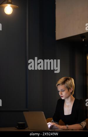 Eine fokussierte professionelle Frau arbeitet fleißig an ihrem Laptop in einem schwach beleuchteten, stilvollen Café und schlürft an einem späten Abend Kaffee. Stockfoto