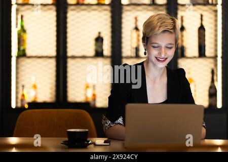 Eine junge professionelle Frau arbeitet an ihrem Laptop in einer eleganten Bar und zeigt Freude und Konzentration bei ihrem Kaffee. Stockfoto