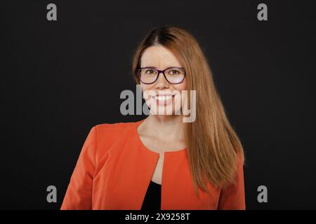 Porträt einer selbstbewussten, professionellen Geschäftsfrau mit Brille und orangefarbenem Blazer, die vor einem schlichten schwarzen Hintergrund lächelt. Stockfoto