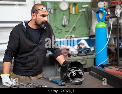 Porträt eines Schmieds während einer Schweißpause. Er beobachtet das Schweißen der beiden Stangen rechts. Auf der Werkbank die Schweißarbeiten h Stockfoto