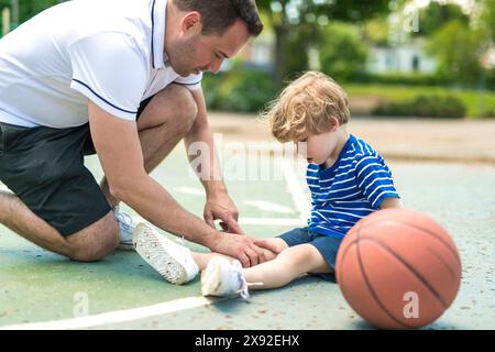 Porträt eines fokussierten erwachsenen Mannes, der einem Jungen mit Knietrauma hilft, nachdem er Basketball auf dem Platz gespielt hat Stockfoto