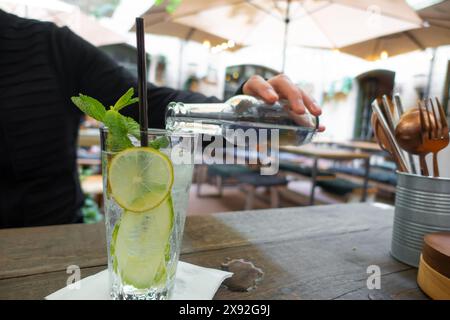 Limonade mit frischer Zitrone auf Holztischhintergrund. Die Hand eines Mannes in einem schwarzen Ärmel gießt ein kohlensäurehaltiges Getränk in ein hohes, durchsichtiges Glas, in dem Stockfoto