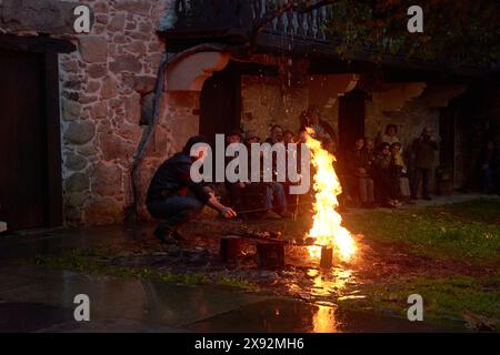 Der Mensch führt ein altes Feuerritual in der Dämmerung in einem malerischen Dorf durch, während eine Gruppe von Zuschauern die faszinierenden Flammen beobachtet Stockfoto