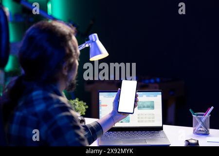 Bärtiger Mann, der ein Mobilgerät vertikal hält und einen isolierten weißen Bildschirm anzeigt. Männlicher Freelancer, der an seinem Laptop arbeitet, während er das Smartphone mit leerer Copyspace-Mockup-Vorlage ergreift. Stockfoto