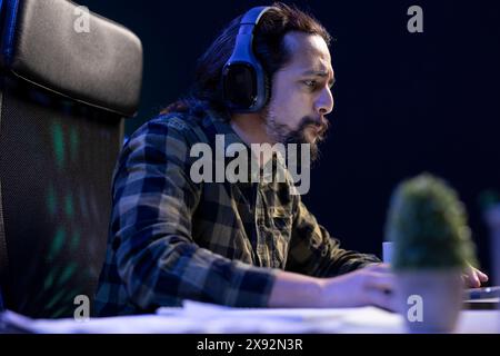 Fokussierter Mann bringt seinen Laptop zu virtuellen Meetings und surft mit schnurlosen Kopfhörern im Internet. Bärtiger Kerl, der Multitasking macht, indem er Filme ansieht und online Hausaufgaben macht. Stockfoto