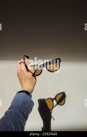 Weibliche Hand, die bei natürlichem Sonnenlicht eine stilvolle Sonnenbrille über die weiße Wand hält Stockfoto