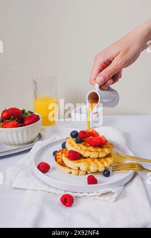 Nahaufnahme der weiblichen Hand, die Ahornsirup aus der Untertasse auf süße, hausgemachte Waffeln mit Beeren gießt Stockfoto