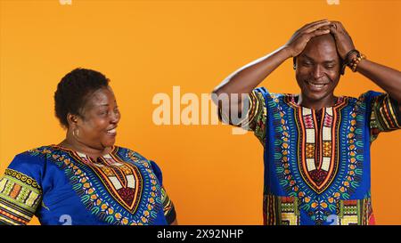 Fröhliche afroamerikanische Leute, die über etwas im Studio lachen, Spaß mit Witzen haben und sich über orangefarbenen Hintergrund amüsieren. Verheiratetes Paar lacht vor der Kamera, dumme Partner. Stockfoto