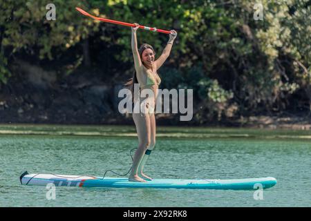 Das lächelnde Latina-Mädchen steht triumphierend auf ihrem Paddleboard und hebt ihr Paddel mitten im See in die Luft. Genieße einen siegreichen Moment Stockfoto