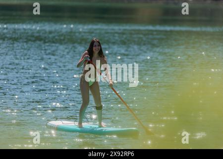 Junge Lateinfrau im Bikini, die auf einem See paddelnd surft, mit Büschen, die den Vordergrund des Fotos verdecken und eine ruhige natürliche Szene schaffen Stockfoto
