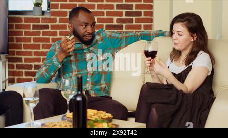 afroamerikanischer Mann flirtet mit Frau auf Couch auf der Apartmentparty und genießt Unterhaltung. Gast, der der Frau zu Hause während des Geburtstags der Freunde die Warteschlangen erzählt und Alkohol trinkt Stockfoto