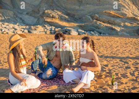 Drei hispanische Freunde sitzen am Sandstrand, spielen auf einer Ukulele und lachen, während sie den wunderschönen Sonnenuntergang genießen. Mit kalten Bieren in der Hand, t Stockfoto