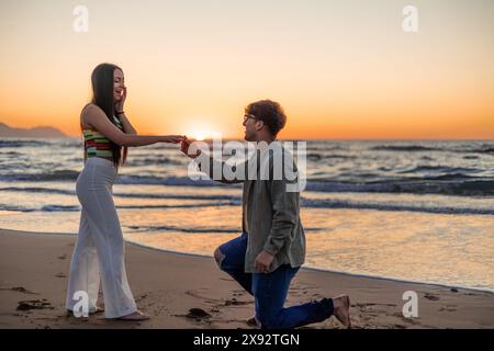 In einem romantischen Sonnenuntergang am Strand kniet sich ein junger Mann nieder und schlägt seiner multikulturellen lateinischen Freundin einen Ring vor, was einen Moment voller W Stockfoto