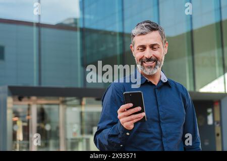 Ein reifer männlicher Arbeiter, der sich eine Finanz-App auf seinem Handy ansieht. Person mittleren Alters, die ein Handy mit einem Lächeln hält. Gutaussehender mittelgroßer Mann, der am Arbeitsplatz mit einem Smartphone lächelt. Hochwertige Fotos Stockfoto