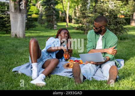 Entspannte schwarze Paare sitzen mit Laptop auf dem Gras auf dem College Campus und essen zwischen den Vorlesungen zu Mittag Stockfoto