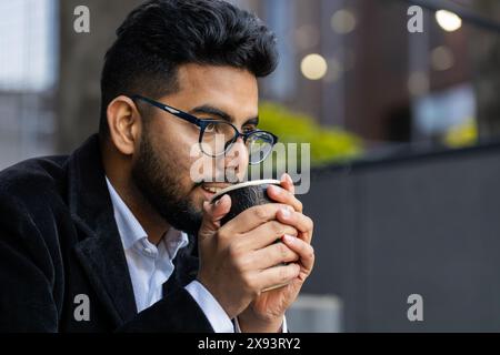 Glücklicher, bärtiger indischer Geschäftsmann, der morgens Kaffee, ein heißes Getränk genießt und draußen lächelt. Entspannen, eine Pause einlegen. Ein arabischer Hindu-Freelancer trinkt Kaffee, um in die Innenstadt zu gehen. Lifestyle Stockfoto