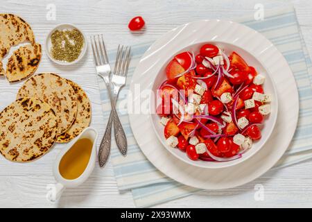 Frischer Tomatensalat mit roten Zwiebeln und weißem salzigem griechischem Käse in Schüssel auf weißem strukturiertem Holztisch mit Gabeln und gegrilltem Flachbrot, horiz Stockfoto